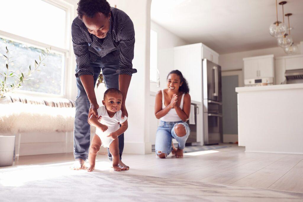 Parents Encouraging Smiling Baby Daughter To Take First Steps And Walk At Home
