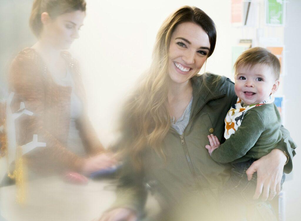 Woman holding a baby standing next to a cash register with shop assistant, smiling at camera.