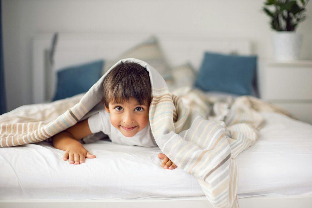 five year old boy covered himself with a blanket on the bed in the white bedroom of the nursery