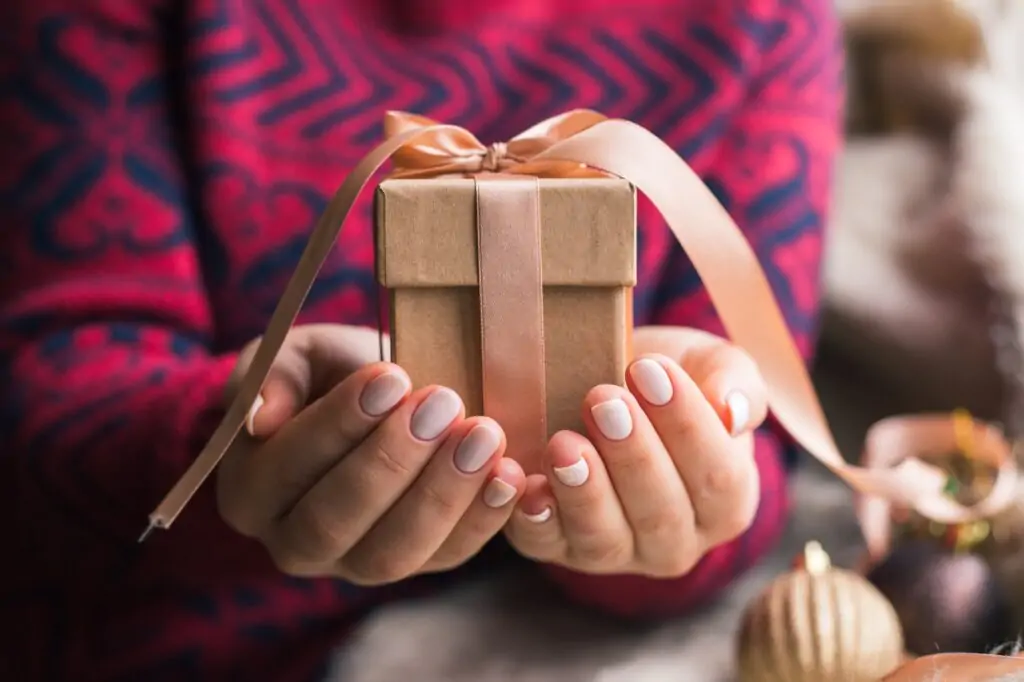 Les meilleurs cadeaux pour maman : idées originales et touchantes A woman holding a Christmas gift with a ribbon on a wooden table. Christmas presents and New Year