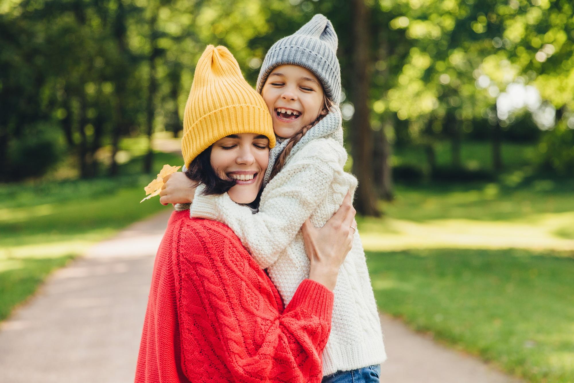 Beautiful female and her daughter have warm embrace, dressed in knitted clothes