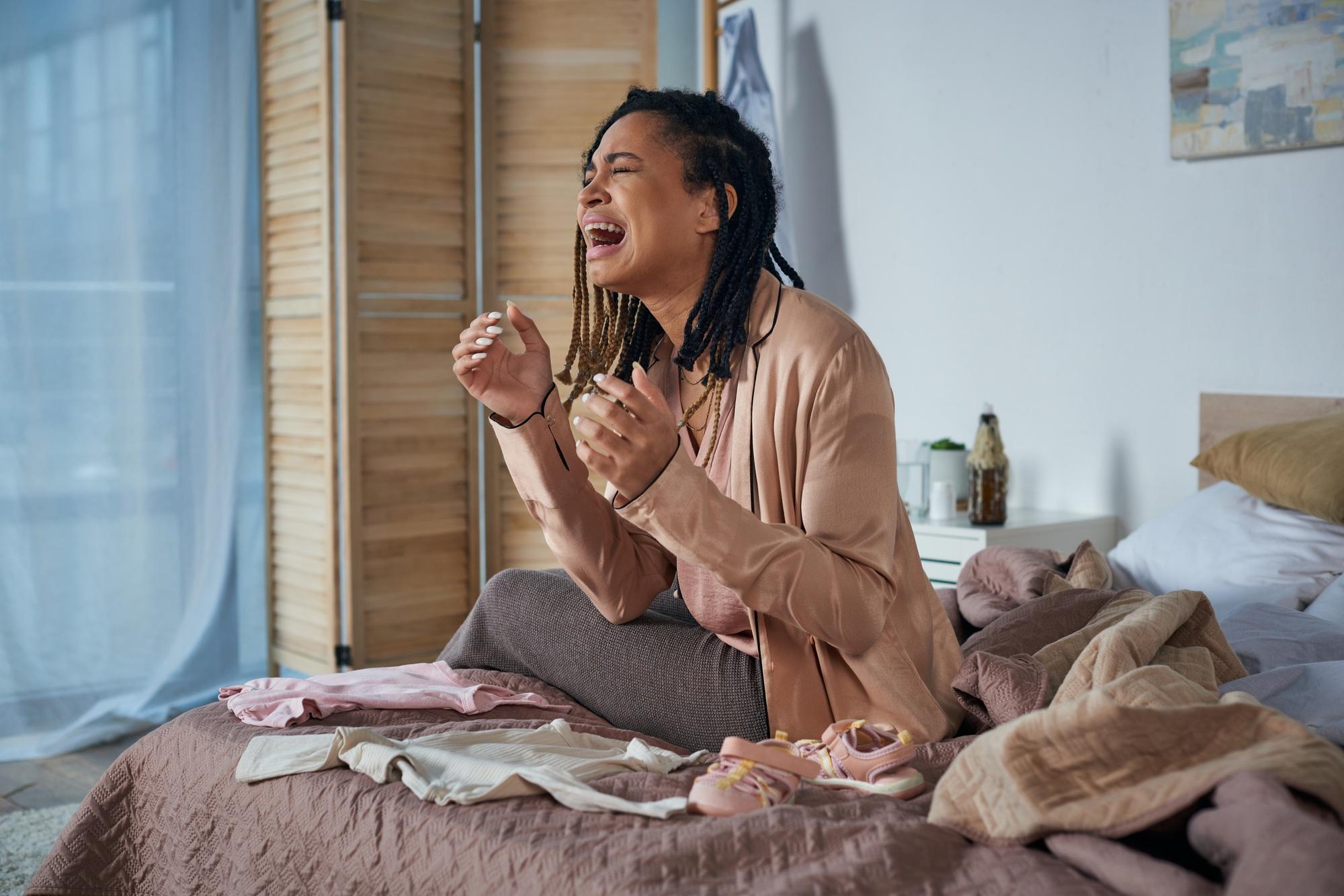 grief, depressed african american woman crying near baby clothes on bed, miscarriage concept