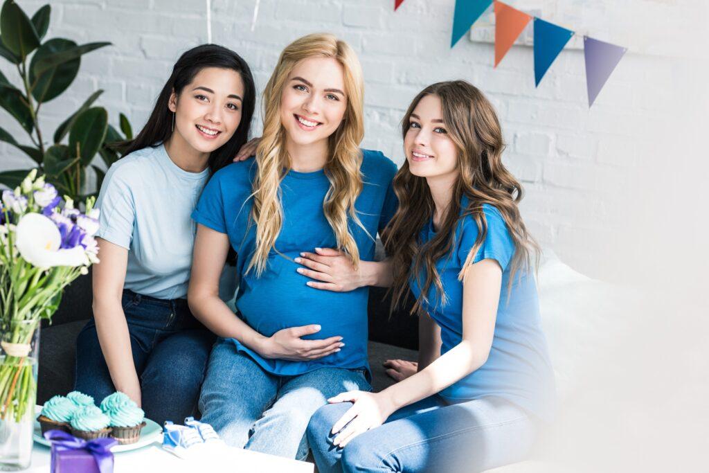 multicultural friends and pregnant woman touching belly and sitting on sofa at baby shower party