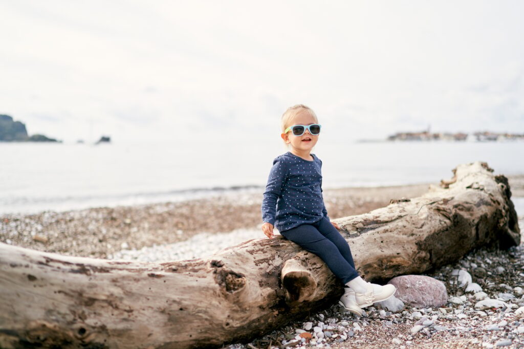 Little girl in sunglasses sits on a snag on the beach