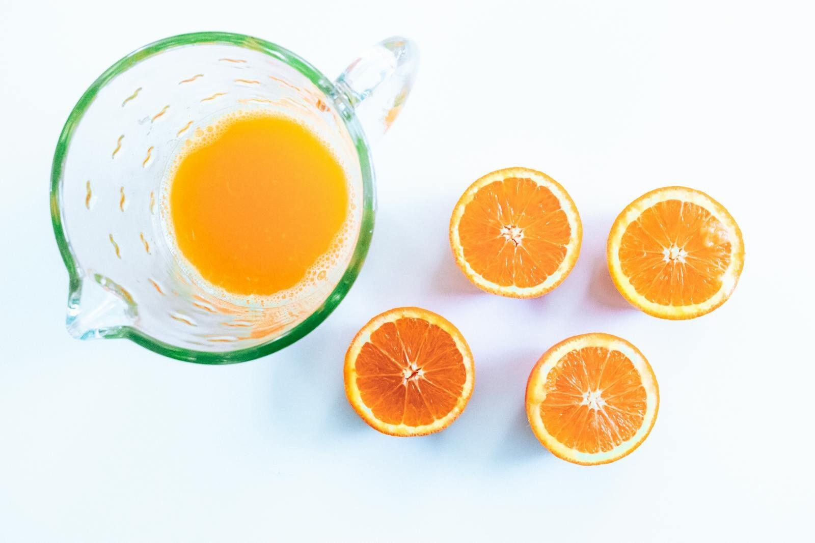 Glass pitcher of fresh orange juice with four halved oranges on a white surface.