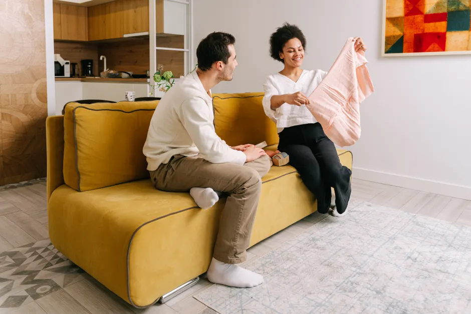 Après bébé, 5 mercis pour le couple, sans match de comptabilité A cheerful couple engaged in conversation on a modern yellow sofa, discussing baby clothing in a stylish living room.