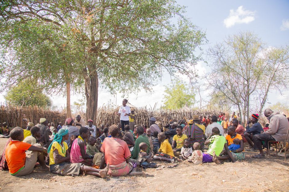 A diverse group of adults and children gather under a tree for a community meeting outdoors.