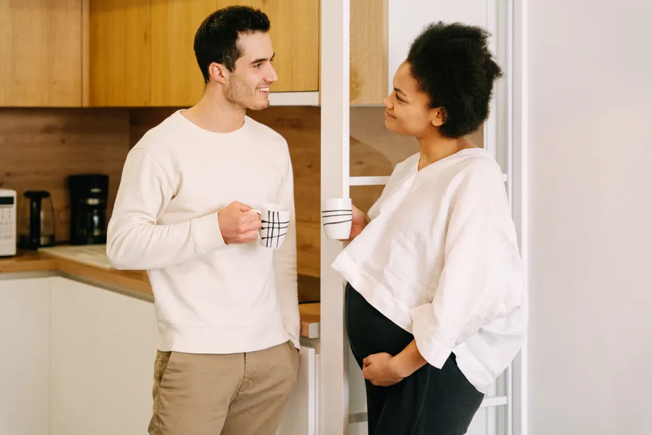 Grossesse : 4 repères pour parler en couple sans perdre le fil A happy expectant couple enjoying a conversation in the kitchen with coffee mugs.
