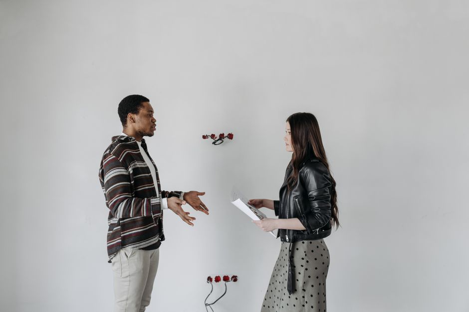 A man and woman in stylish outfits having a discussion in an interior setting with a white wall.