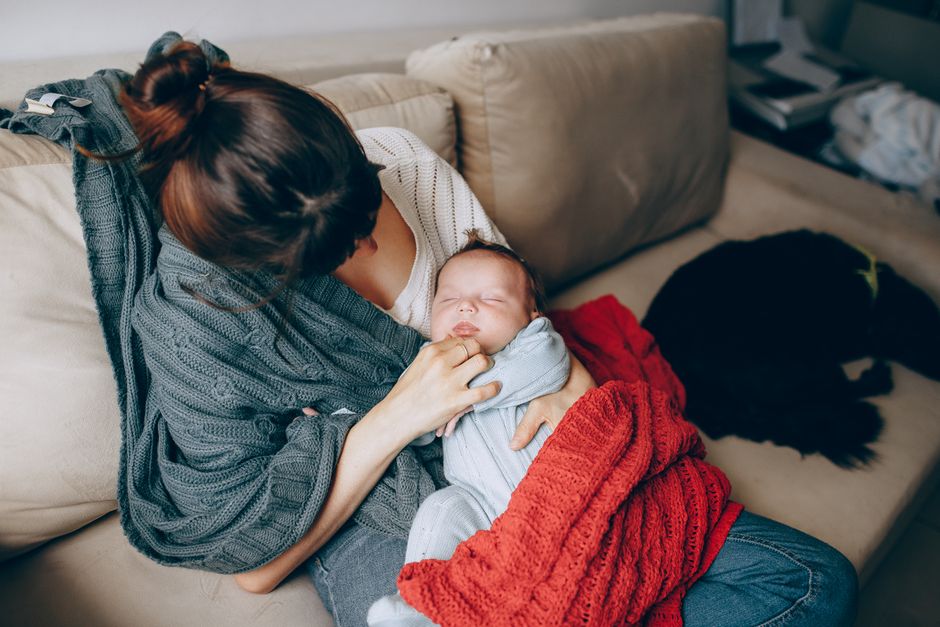 Après bébé, 5 repères pour refaire une place à l’intimité A tender moment of a mother comforting her sleeping baby while resting on a sofa indoors.