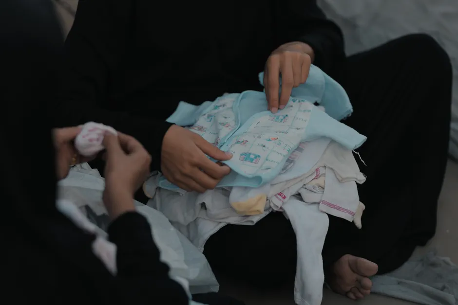 A woman in a Gaza refugee camp sorts a pile of baby clothes, highlighting daily life challenges.