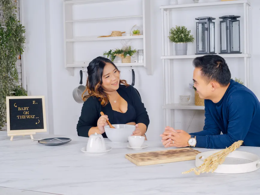 Couple après bébé : 5 rituels pour retrouver le « nous » Asian couple in a kitchen setting with a pregnancy announcement sign 'Baby on the Way', smiling at each other.