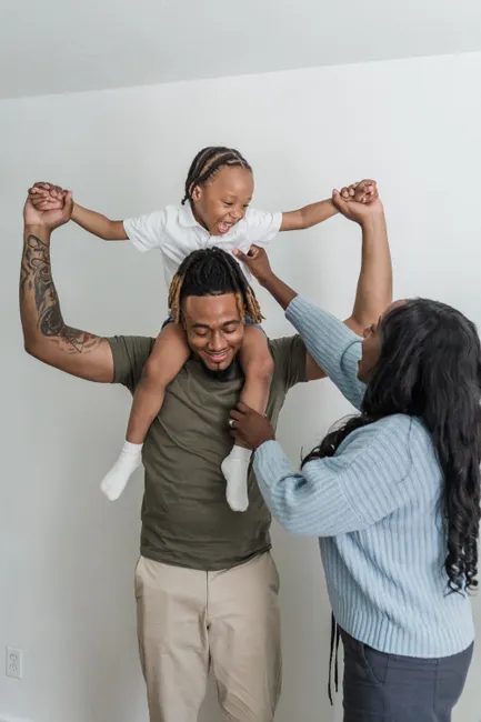 Father joyfully lifts his smiling child on shoulders, supported by mother indoors.