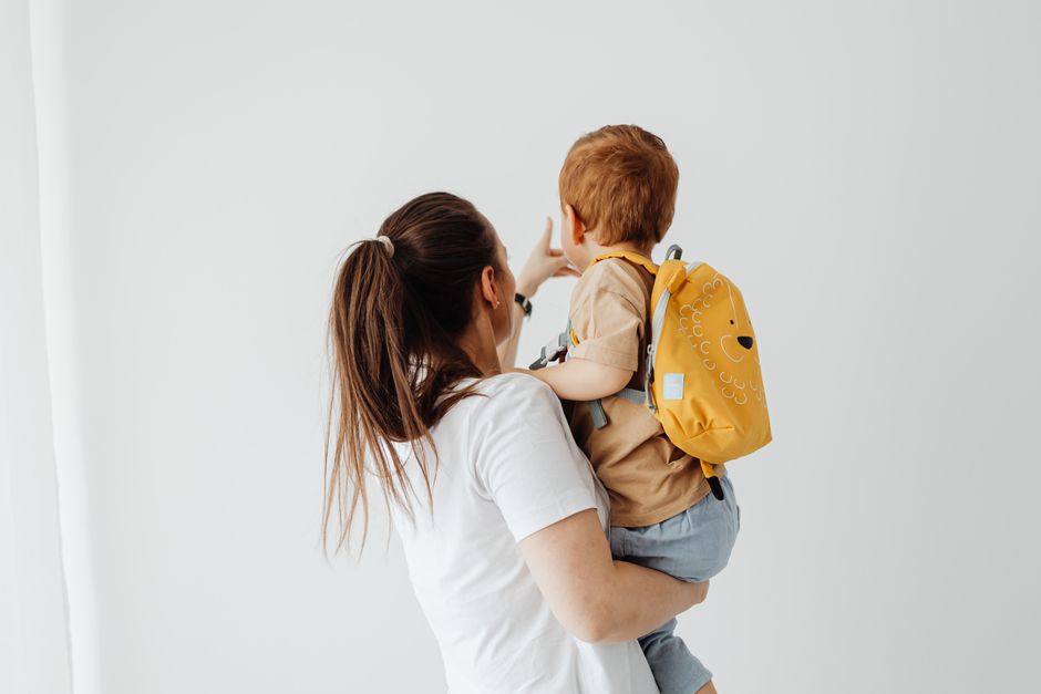 Après bébé : 7 scènes banales qui parlent du couple, apparemment Mother holding her son with a yellow backpack indoors, showcasing family togetherness.