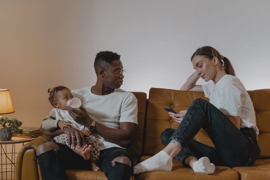 Couple après bébé : 5 gestes pour garder le lien malgré les nuits hachées Parents relaxing on a sofa while feeding their baby, enjoying family time with a cellphone.