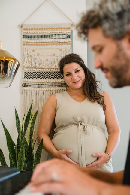 Smiling young pregnant female in beige dress listening to bearded husband playing piano in room