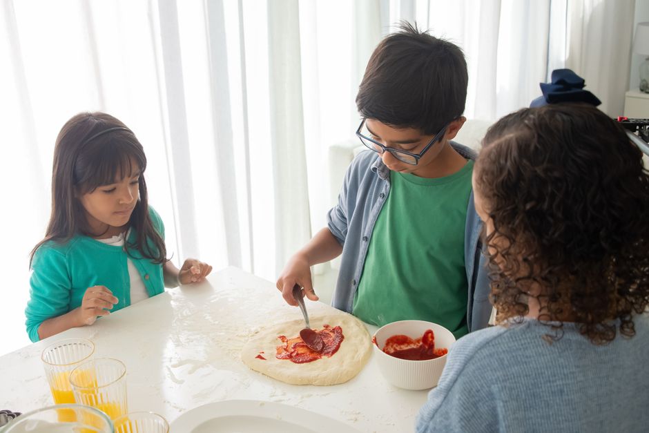 Temps de qualité en famille : 6 idées pour les 4 à 16 ans, même quand ça déborde Three children are joyfully making pizza with tomato sauce in a bright indoor kitchen.