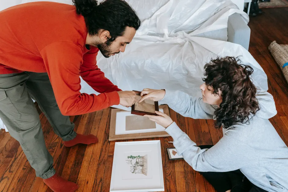 Décorer en couple : 7 règles pour éviter la guerre du canapé A couple arranges picture frames in a cozy living room, planning their decor.