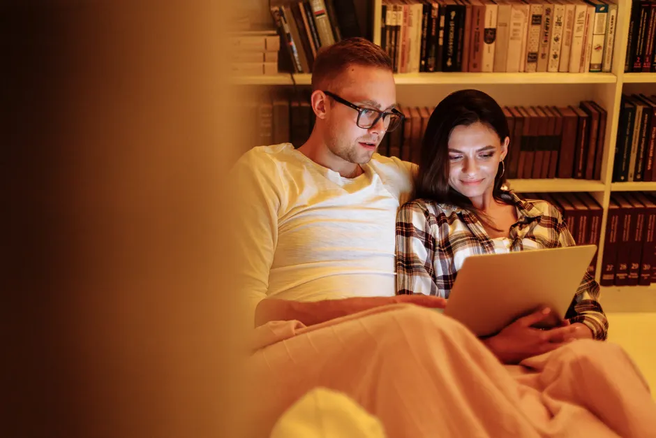 Couple en pilote automatique : 5 gestes pour retrouver le lien sans grand soir A couple enjoys a cozy evening reading together on a tablet in a home library.