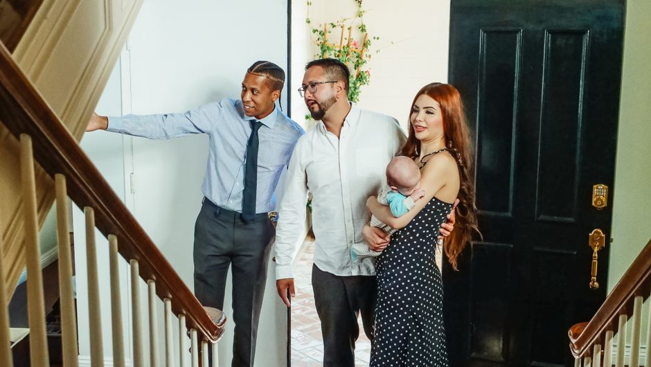 A family with a baby tours a new home with a realtor, exploring indoor spaces and possibilities.