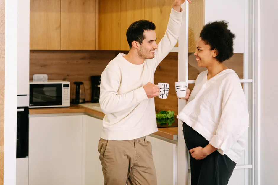 Après bébé, 7 gestes pour retrouver son couple sans jouer à Love Actually A happy interracial couple shares a coffee break in the kitchen, celebrating pregnancy moments.
