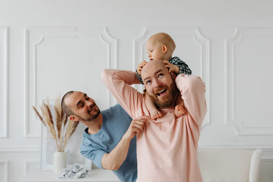 Humour dans le couple après bébé : 6 règles pour rire sans se piquer A heartwarming scene of a baby enjoying playful time with loving same-sex parents indoors.