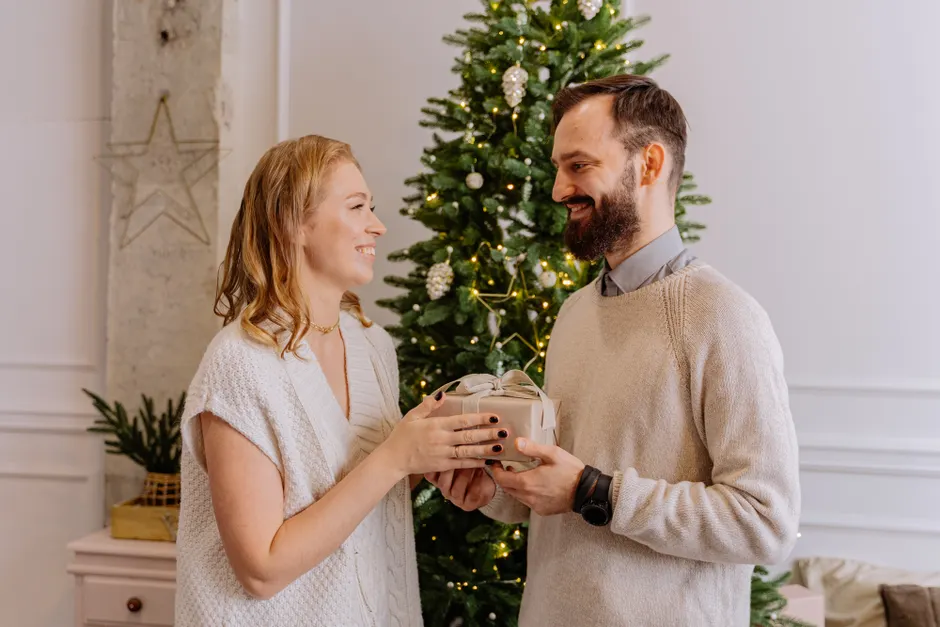 Motivation extrinsèque dans le couple : 5 gestes sans dette affective A smiling couple exchanges a gift in front of a decorated Christmas tree, adding warmth and joy to the holiday season.