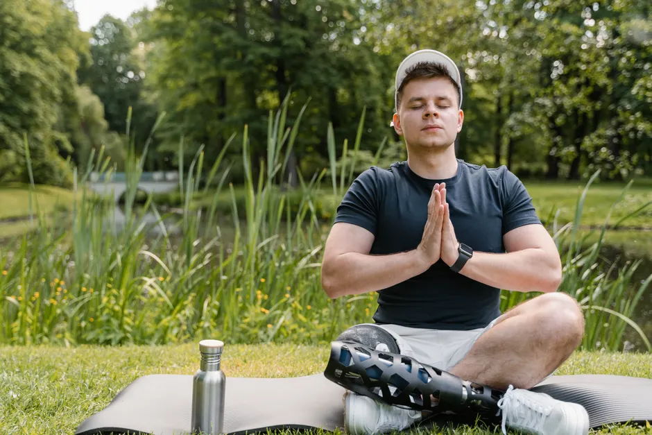 Adult male with prosthetic leg meditates on yoga mat in park, promoting wellness and relaxation.