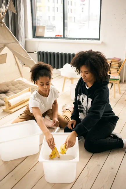 African American mother and daughter putting banana peel into white container while sorting garbage on wooden floor in light room with blanket fort