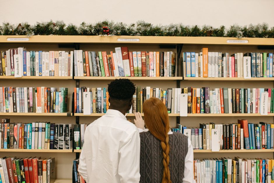 Livre pour couple : 5 critères avant de tourner les pages An interracial couple browsing books on a bookshelf decorated with holiday garlands.