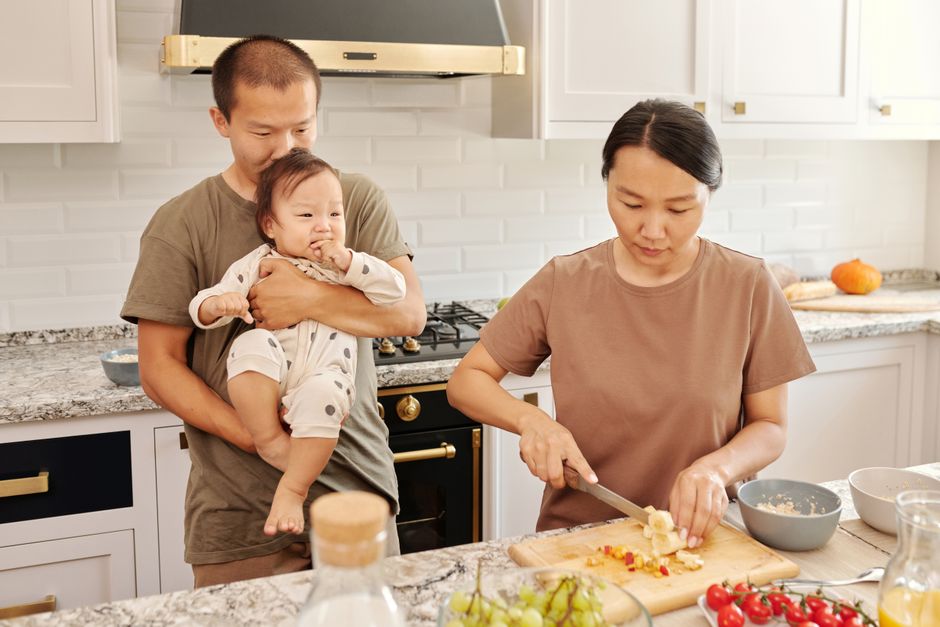 6 loisirs à deux après bébé quand le temps manque Asian parents with toddler cooking together, sharing a moment in the kitchen.