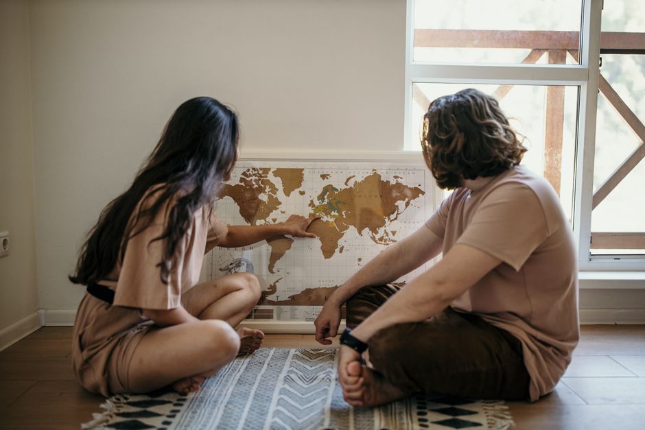 Premier voyage en couple : le crash-test amoureux en 7 règles Couple sitting on floor pointing at a world map, planning their next travel adventure indoors.