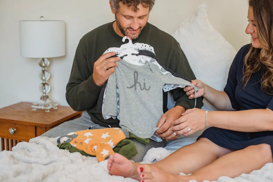 Après bébé, 6 phrases pour s’écouter en couple sans mode d’emploi Crop smiling barefoot expectant female with male beloved watching baby clothes on soft bed in house