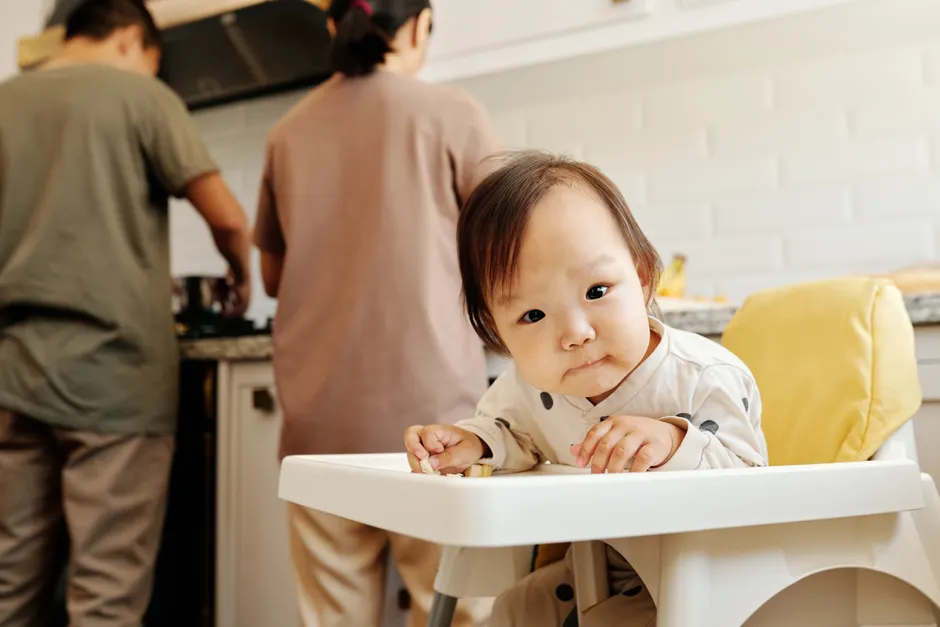Cute toddler sitting in a high chair while parents prepare food in the kitchen.