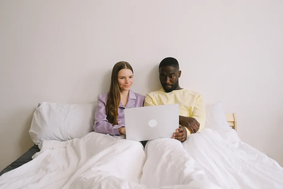 Sexualité après bébé : 58 % sous le seuil FSFI à 6-12 mois Interracial couple sitting in bed using a laptop, showcasing love and technology at home.