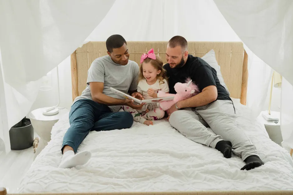 Interracial gay couple reading a book with their child on a bed, symbolizing family love and togetherness.