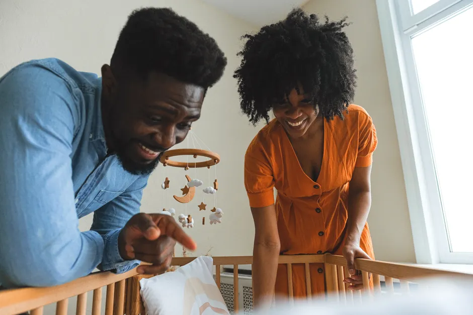 Après bébé, 4 gestes pour entretenir le lien du couple Joyful African couple smiling at their baby's crib under a cozy indoor setting.