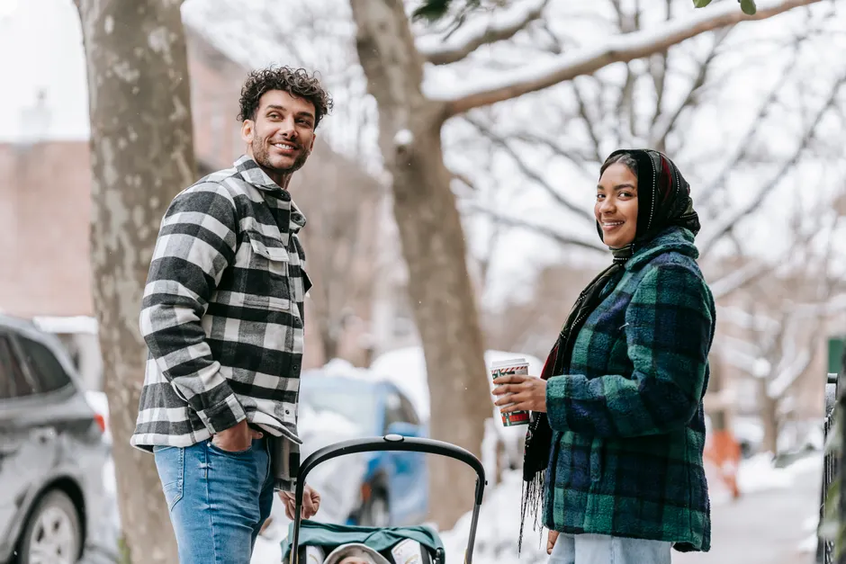 Après bébé, 5 gestes pour retrouver l’amitié dans le couple Low angle side view of positive ethnic mother with cup of takeaway coffee standing on street with baby in stroller and father and looking away