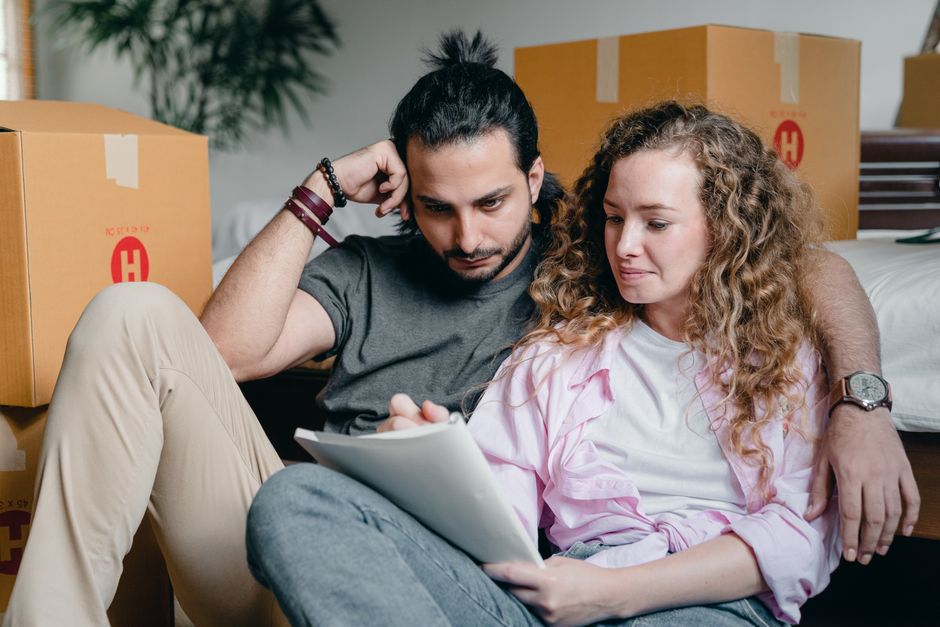 Vision de couple : 5 questions pour avancer ensemble sans réunion de crise Pensive male and female in casual clothes sitting together among boxes and writing notes in notebook while leaning on bed