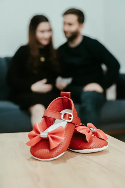 Red baby shoes on table with blurred expectant couple in background, symbolizing anticipation and joy.