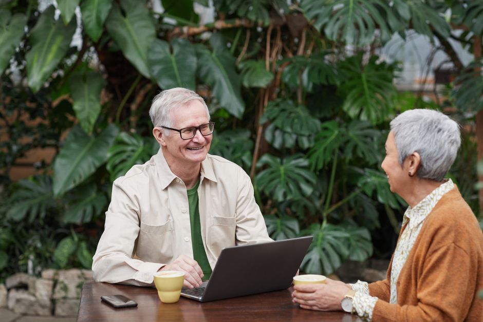Senior couple smiling and enjoying coffee and conversation with laptop in a lush garden.