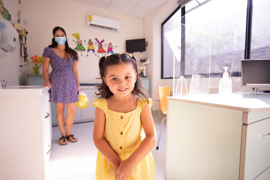 Consultation d’enfant : 5 réflexes pour parler clair A cheerful child in a yellow dress at a pediatric clinic with doctor in background.