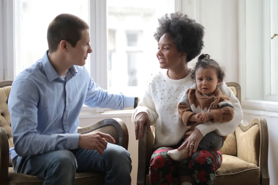 Avant 3 ans, parler sans pression quand le stress s’invite Diverse mother and father talking to each other while sitting in cozy armchairs with upset toddler