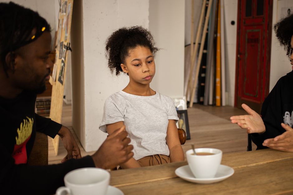 Communication dans le couple : 5 réglages quand la charge mentale déborde Sad African American girl sitting at wooden table with cups of coffee near black parents having conversation in light room